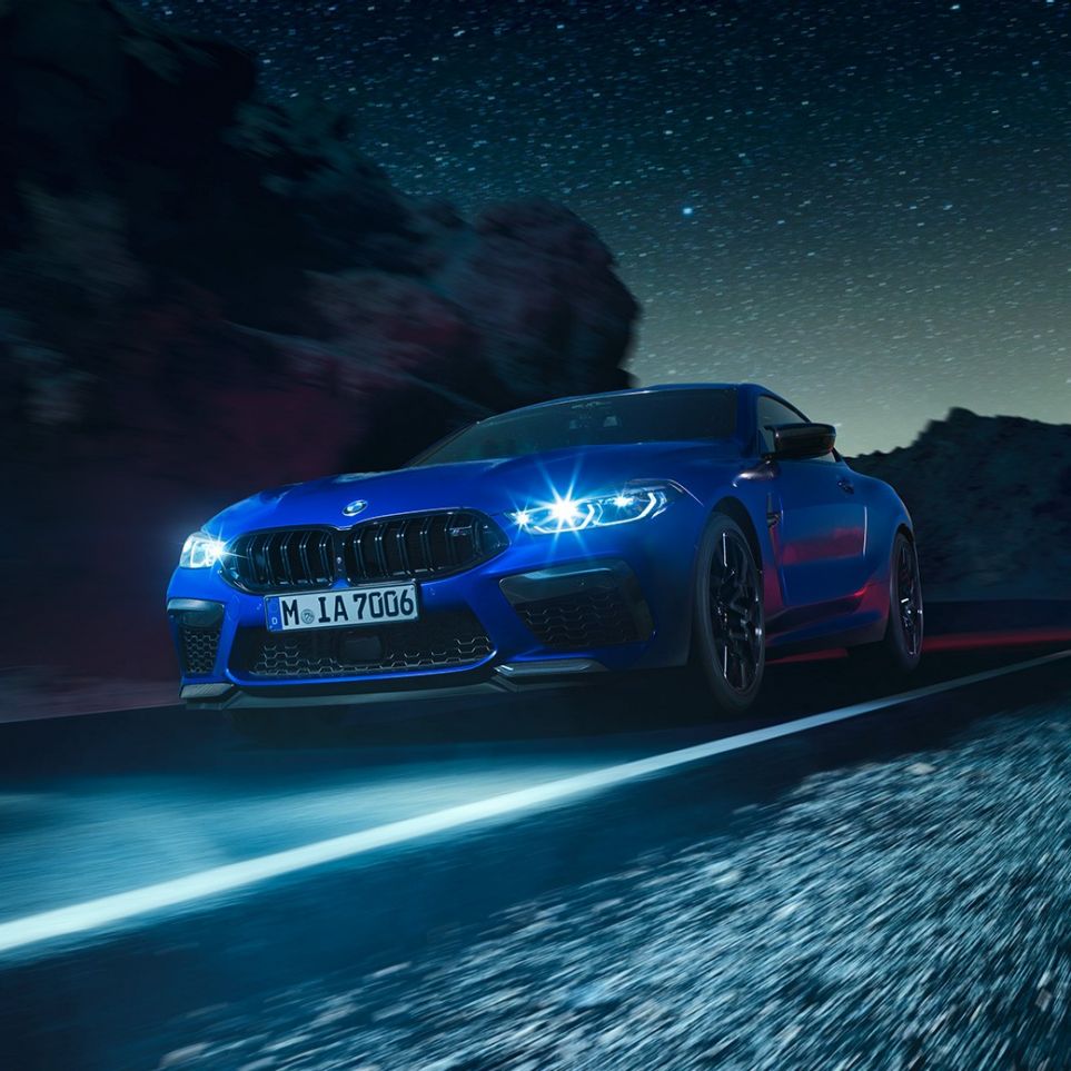 Fahrendes BMW M8 Competition Coupé, Dreiviertel-Frontansicht. F92 BMW M8 Competition Coupé in Marina Bay Blau metallic mit strahlenden LED-Scheinwerfern, Dreiviertel-Frontansicht, fahrend bei Nacht. F92