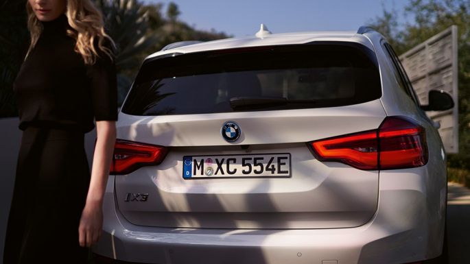Heck mit BMW i spezifischem Diffusor BMW iX3 SUV G08 Heckansicht mit Frau Heck mit BMW i spezifischem Diffusor BMW iX3 SUV G08 2020 Mineralweiß metallic Heckansicht mit Frau