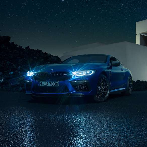 BMW M8 Competition Coupé, vue de trois quarts avant F93. BMW M8 Competition Coupé en Marina Bay Blau metallic avec phares LED allumés, vue de trois quarts avant à l’arrêt devant une villa, la nuit F93.