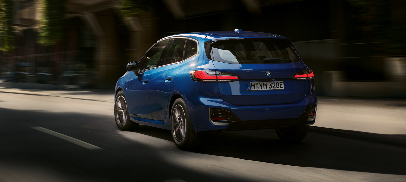 BMW Série 2 Active Tourer 230e Hybride Rechargeable U06 2021 sur une route en ville BMW Série 2 Active Tourer 230e Hybride Rechargeable U06 2021 M Portimao Blau métallisé sur une route en ville, vue de trois quarts arrière