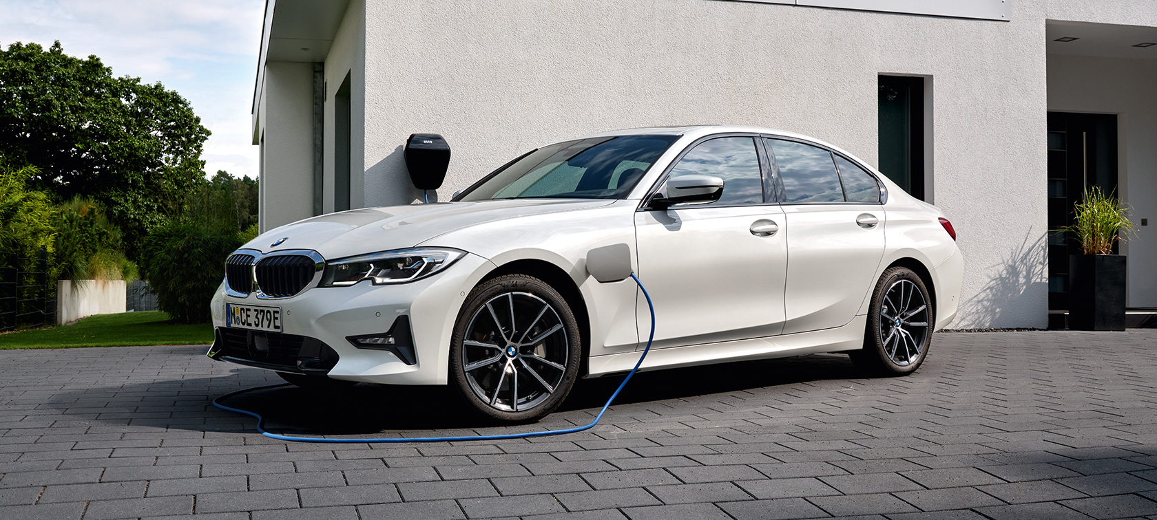 BMW Série 3 Berline hybride rechargeable 2021 G20, vue de trois quarts de profil branchée à une borne de recharge BMW Série 3 Berline hybride rechargeable 2021 G20, vue de trois quarts de profil branchée à une borne de recharge devant un garage