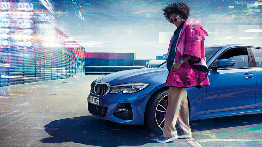 Seitliche dreiviertel Standaufnahme der BMW 3er Limousineauf offenem Parkdeck hinter Frau vor urbaner Kulisse.