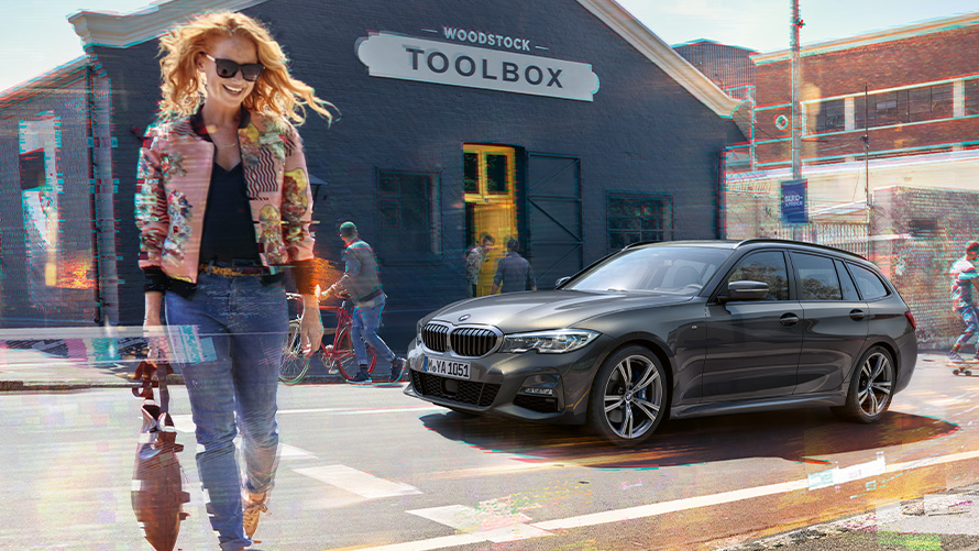 BMW 3er Touring (G21), Dreiviertel-Frontaufnahme auf Straße mit Frau vor Holzwerkzeugladen Frau quert Straße vor BMW 3er Touring (G21) und Holzwerkzeugladen.