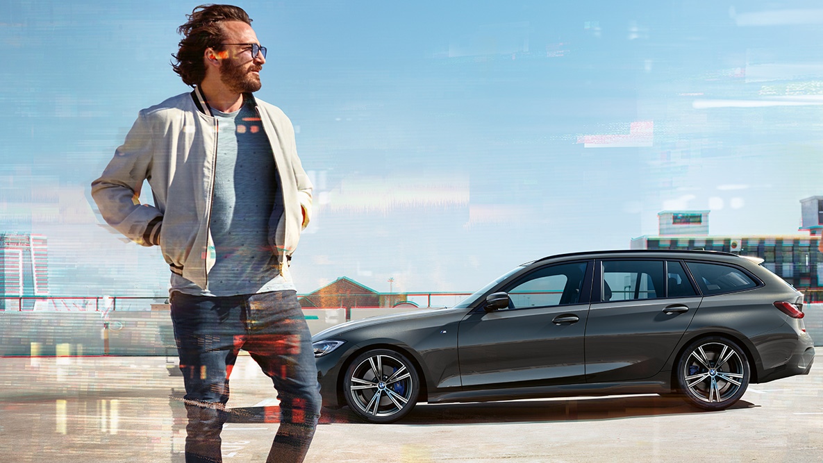 BMW 3er Touring (G21), Dreiviertel-Frontaufnahme Design BMW 3er Touring (G21) Dravitgrau, Dreiviertel-Frontaufnahme im Fahren auf Brücke vor ländlicher Umgebung