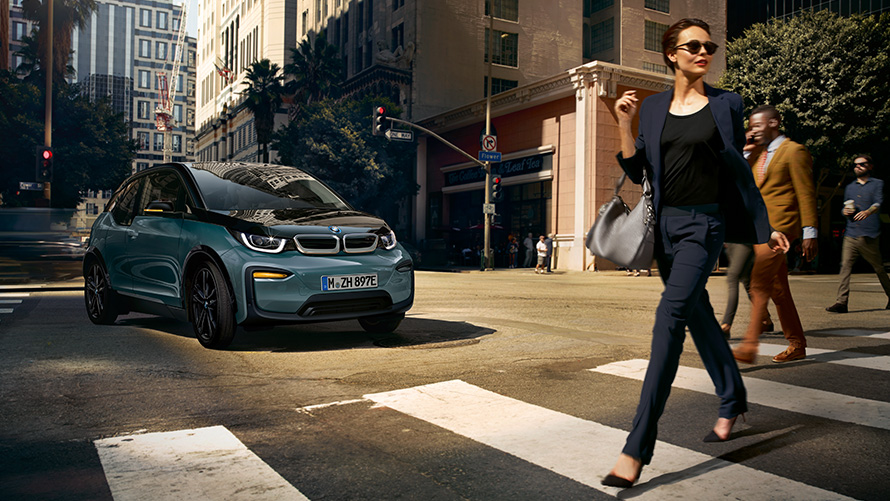 BMW i3 I01 Dreiviertel-Frontansicht mit Frau über Zebrastreifen laufend BMW i3 I01 Blue Ridge Mountain metallic mit Akzent Frozen Grey metallic Dreiviertel-Frontansicht mit Frau über Zebrastreifen laufend