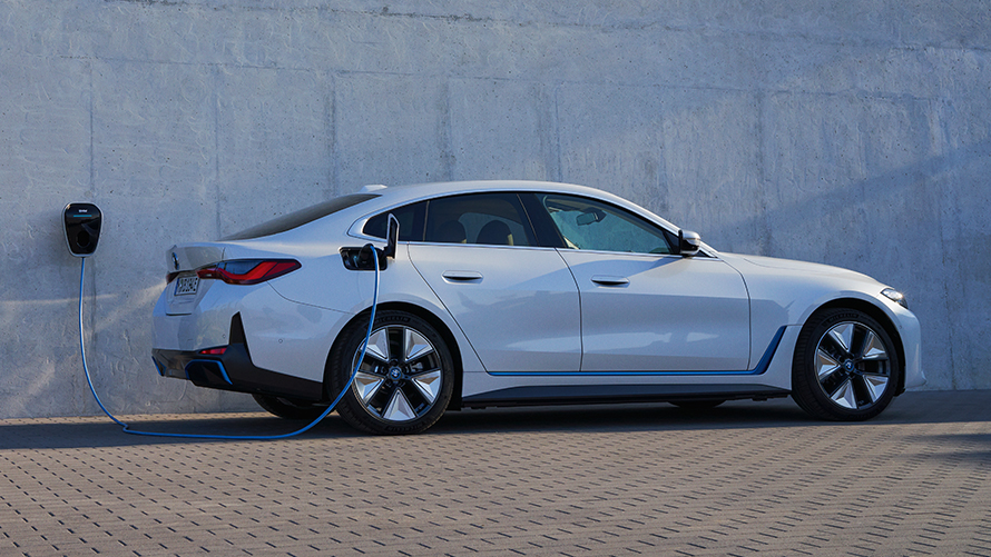 Vue de profil de la BMW i4 eDrive40 G26 2021 avec BMW Wallbox BMW i4 eDrive40 G26 2021 Mineralweiß Vue de profil devant un mur avec une borne de recharge BMW Wallbox