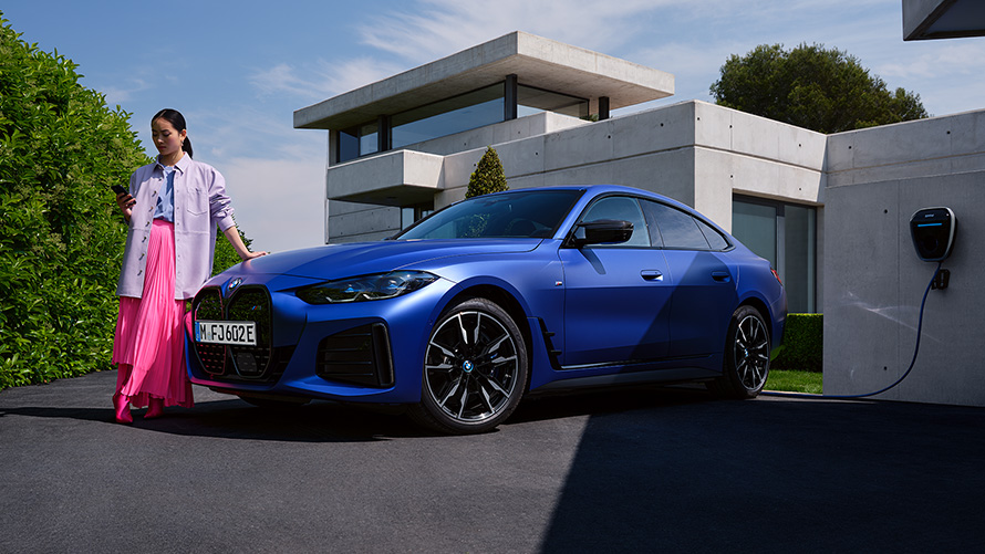 Vue de trois quarts avant de la BMW i4 M50 G26 2021 BMW i4 M50 G26 2021 BMW Individual Frozen Portimao Blau metallic, vue de trois quarts avant avec femme devant une maison