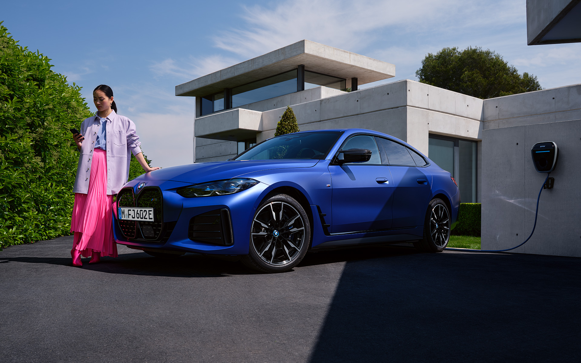 BMW i4 M50 G26 2021 BMW Individual Frozen Portimao Blau metallic Dreiviertel-Frontansicht stehend mit Frau vor Haus