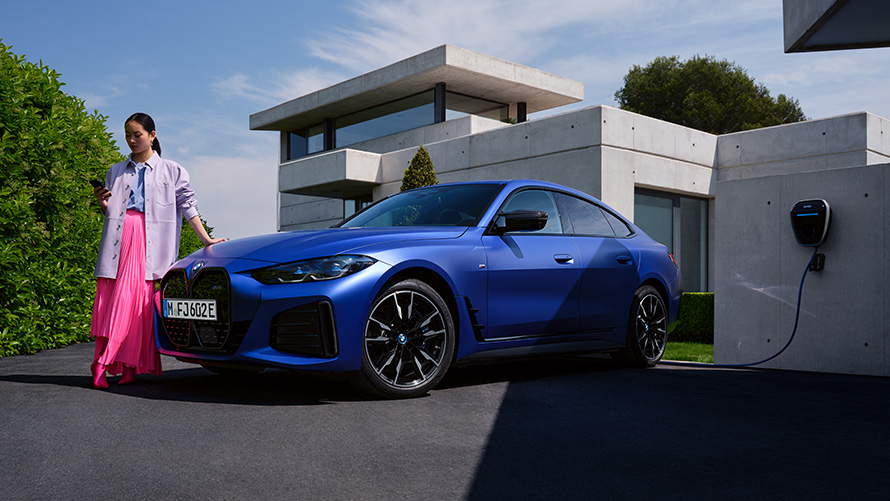 Vue de trois quarts de profil BMW i4 M50 G26 2021 avec femme devant une maison BMW i4 M50 G26 2021 BMW Individual Frozen Portimao Blau metallic, vue de trois quarts de profil avec femme devant une maison