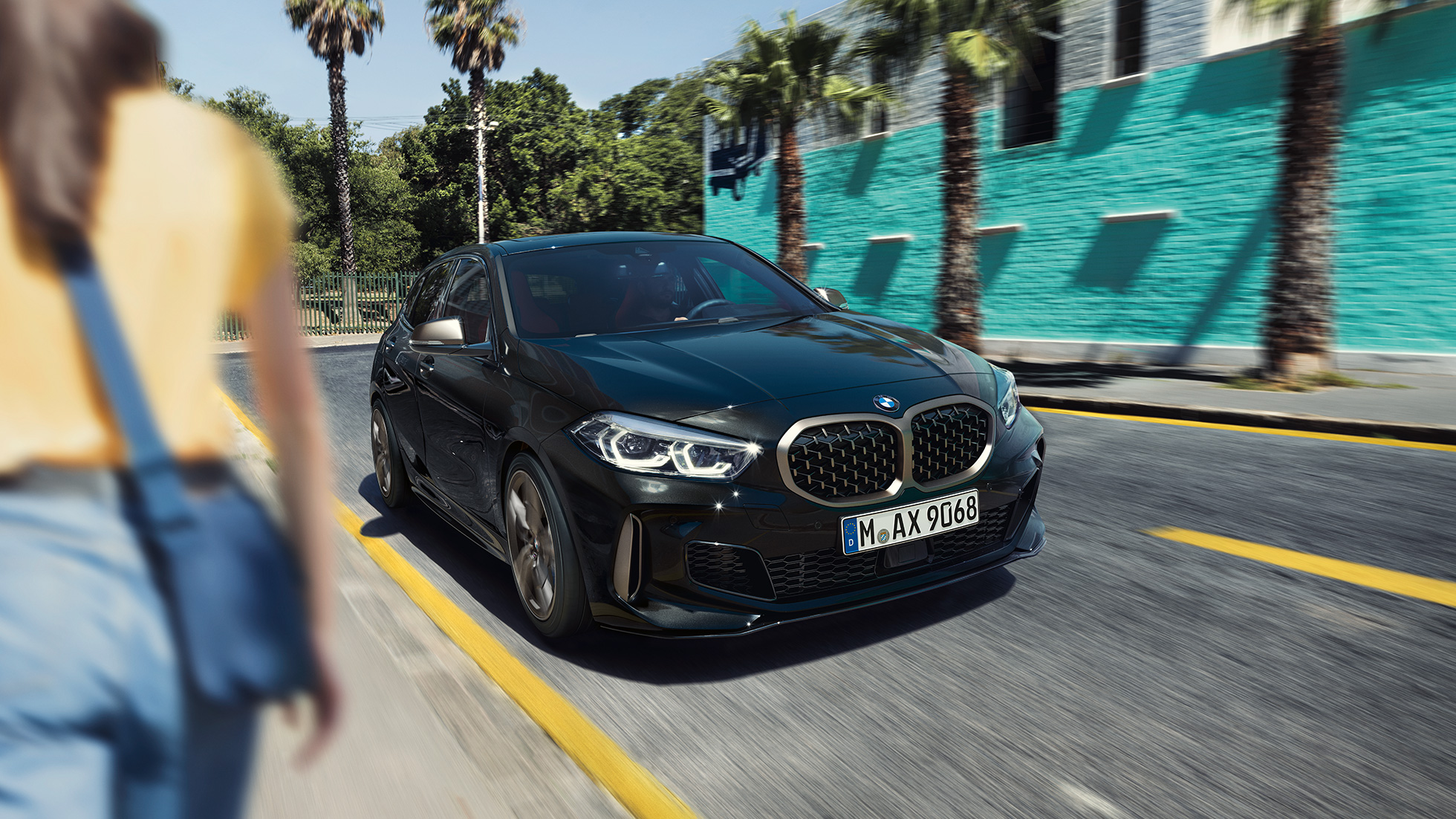 BMW M135i xDrive, Dreiviertel-Frontansicht F40. BMW M135i xDrive in Saphirschwarz metallic, Dreiviertel-Frontansicht fahrend mit Frau im Vordergrund F40.