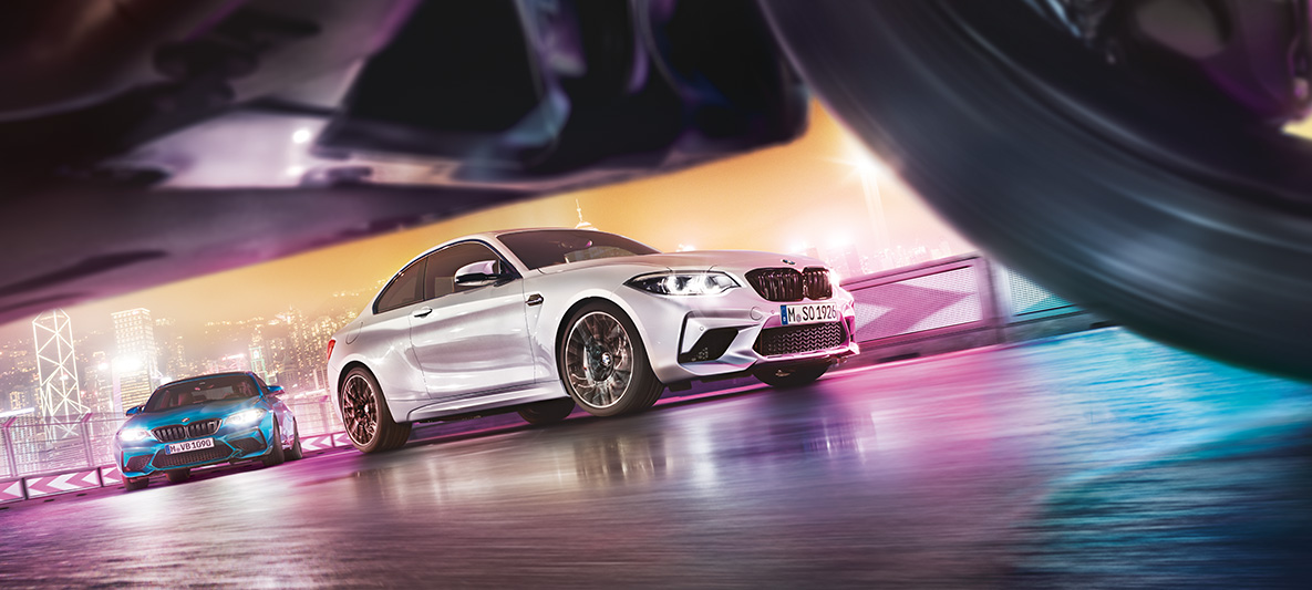 BMW M2 Competition sur du bitume réfléchissant Deux BMW M2 Competition sur le circuit de course, dans le décor nocturne d'une grande ville, vue depuis le sol.