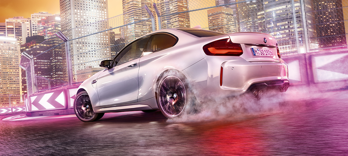 La BMW M2 Competition accélère Une BMW M2 Competition blanche driftant sr le circuit de course, devant une skyline nocturne.