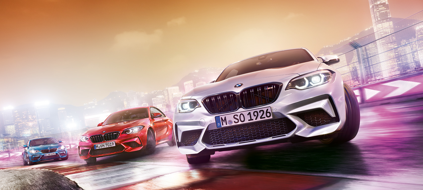 BMW M2 Competition en action Trois BMW M2 Competition font des dérapages sur un circuit de course, dans le décor nocturne d'une grande ville.