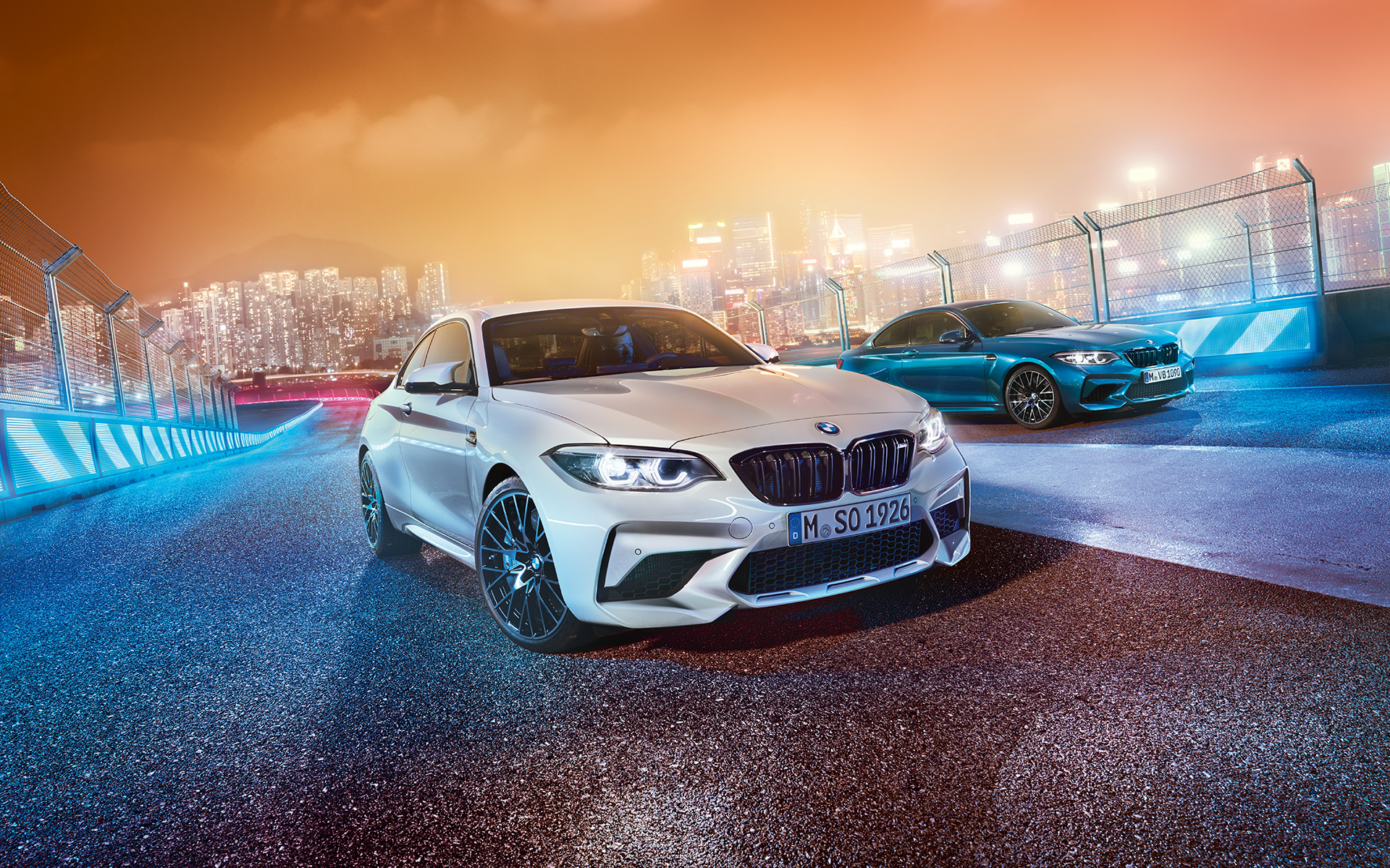 Deux BMW M2 Competition sur le circuit de course Une BMW M2 Competition blanche et une autre bleue, vue oblique de l'avant, dans le décor nocturne d'une grande ville.