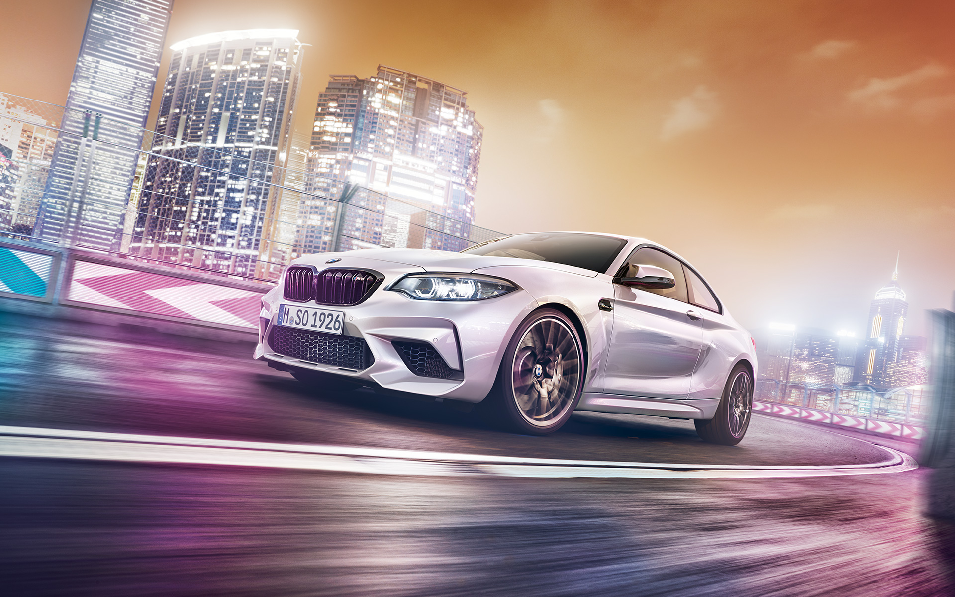 Une BMW M2 Competition blanche avec skyline Une BMW M2 Competition blanche file sur le circuit de course devant une skyline nocturne.