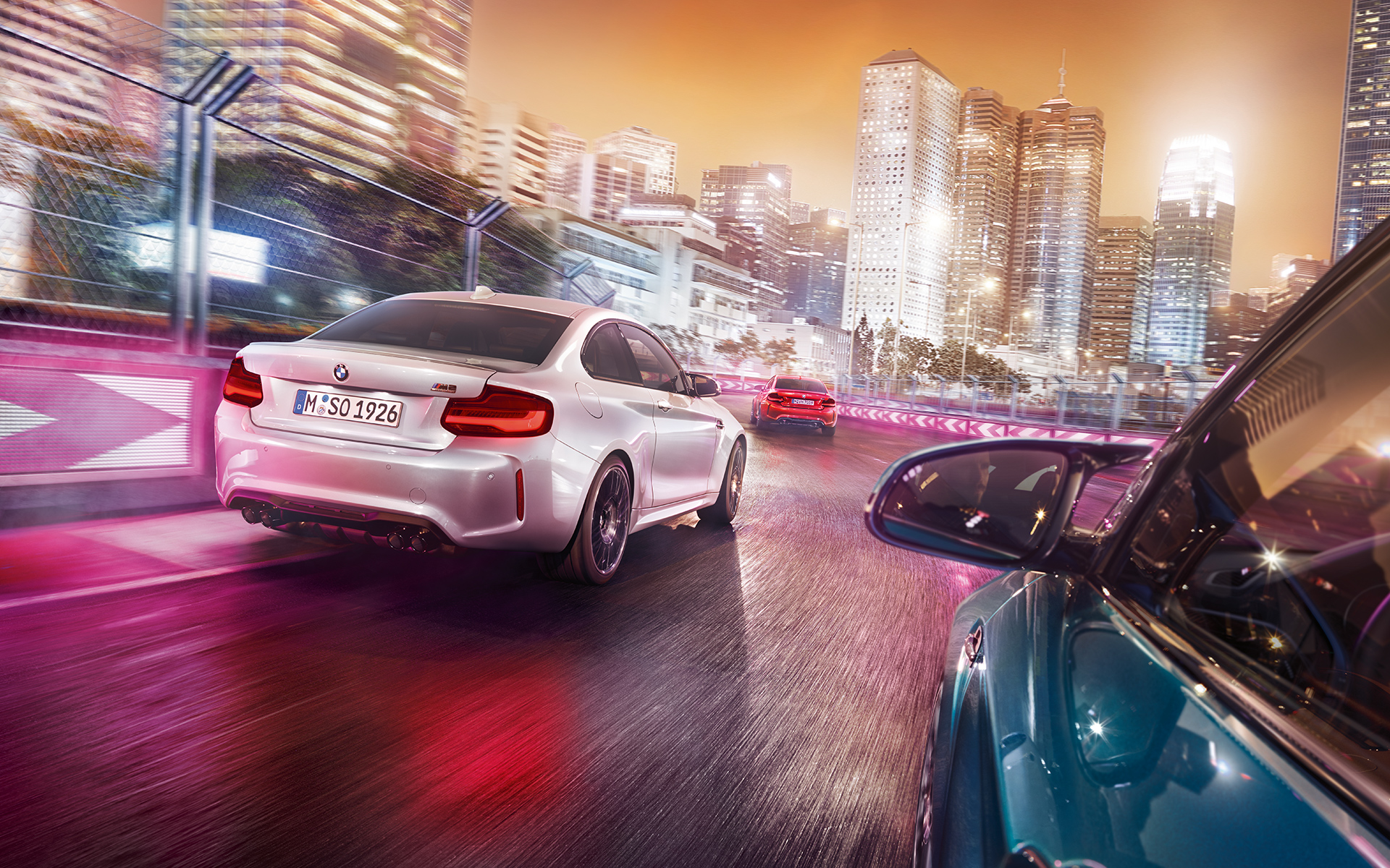 Vue arrière de la BMW M2 Competition Trois BMW M2 Competition vue de l'arrière roulent sur un circuit de course, devant une skyline éclairée.