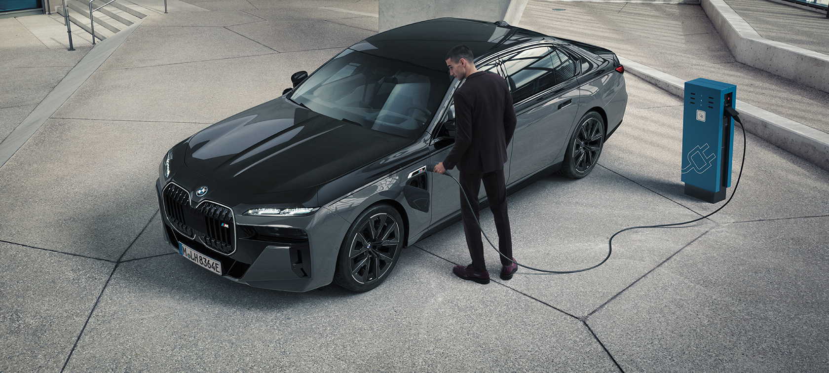 BMW M760e xDrive Limousine G70 M Performance Paket BMW M760e xDrive Limousine G70 M Performance Paket BMW BMW Individual Two-Tone Lackierung Saphirschwarz metallic BMW Individual Dravitgrau metallic Dreiviertel Frontansicht Mann mit Ladekabel neben Ladestation