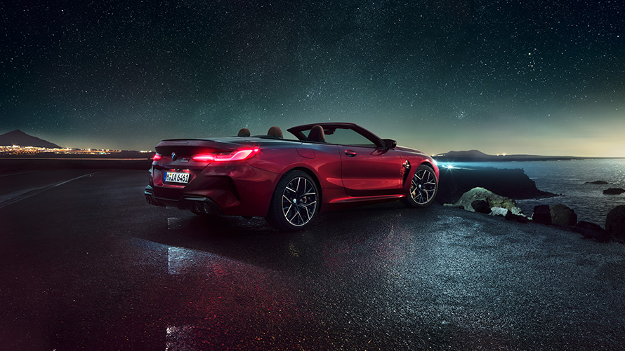 BMW M8 Competition Cabriolet in Motegi Rot metallic mit geöffnetem Verdeck, Dreiviertel-Seitenansicht von hinten, parkend bei Nacht vor Küste.
