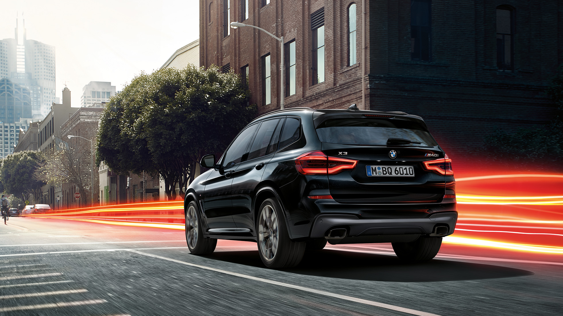 BMW X3 M40i, vue de trois quarts arrière. Extérieur de la BMW X3 M40i en Saphierschwarz metallic, en mouvement, vue de trois quarts arrière devant un paysage urbain avec bandes lumineuses.