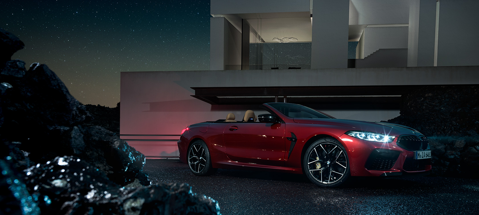 BMW M8 Competition Cabrio, vue de trois quarts de profil. BMW M8 Competition Cabrio en Motegi Rot metallic, vue de trois quarts de profil, à l’arrêt de nuit devant une Villa.