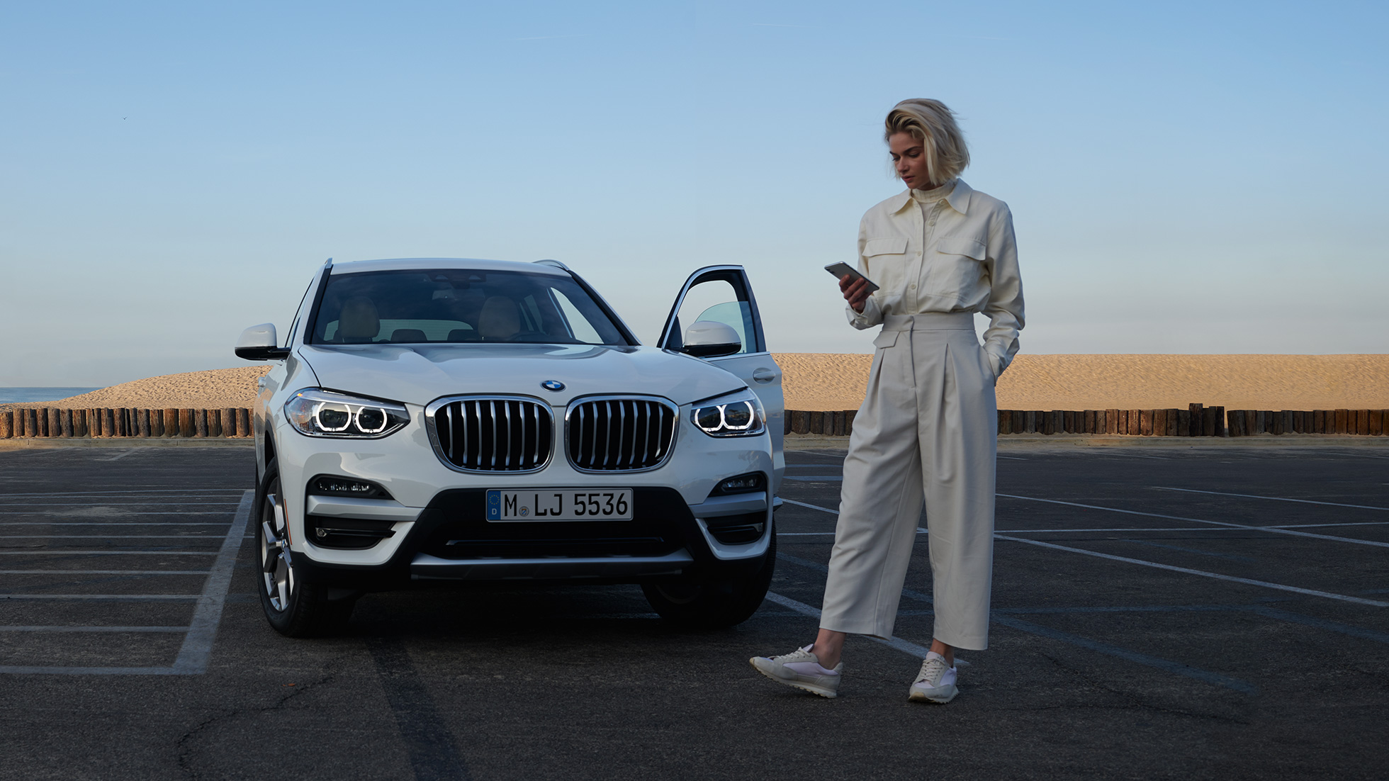 Une femme utilise My BMW App sur smartphone Une femme utilise My BMW App de BMW ConnectedDrive sur son smartphone, devant une BMW X5 blanche.