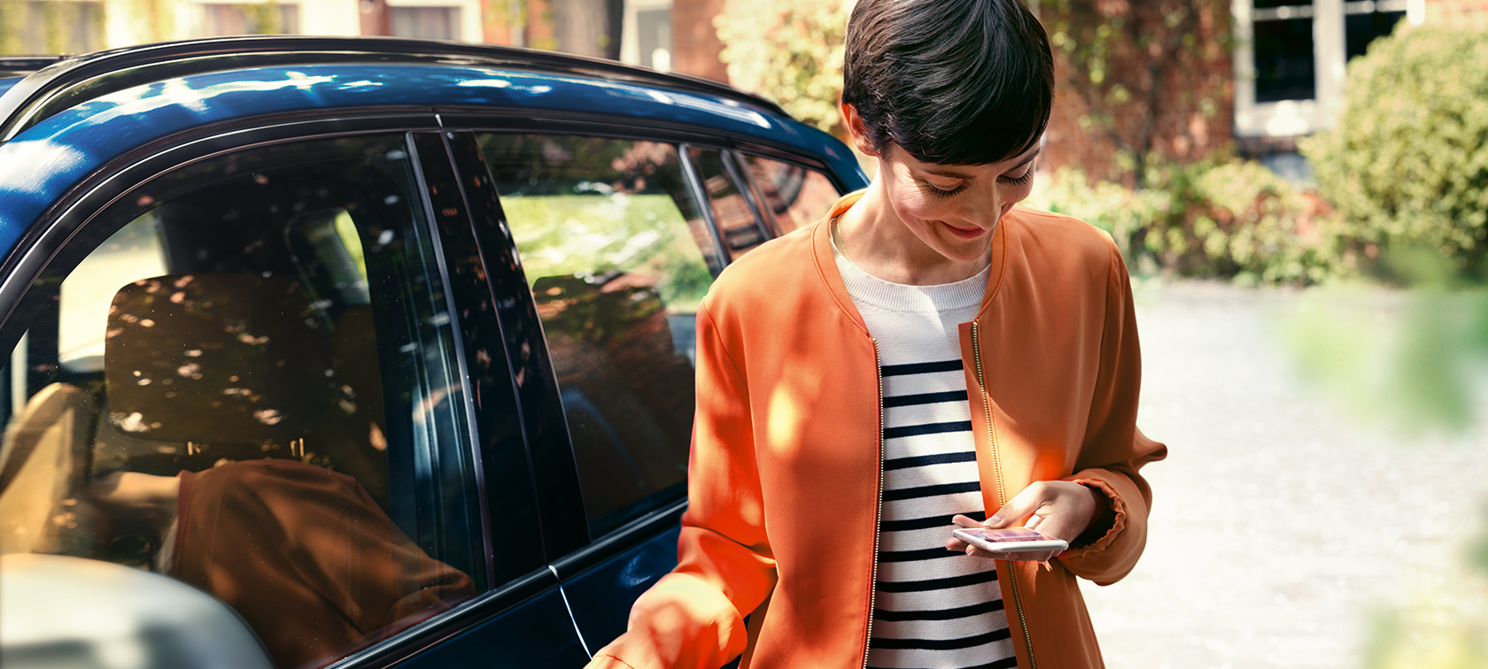 Dunkelhaarige Frau vor BMW Dunkelhaarige Frau in orangener Jacke vor BMW schaut auf ihr Smartphone