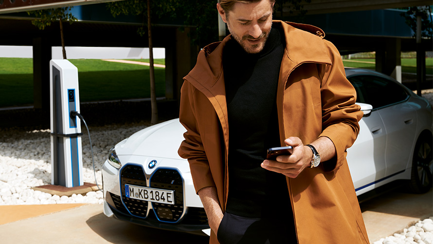BMW Service Inclusive Dauer und Laufzeiten für elektrische Fahrzeuge BMW Service Inclusive Mann schaut auf Smartphone vor BMW i4 an Ladesäule