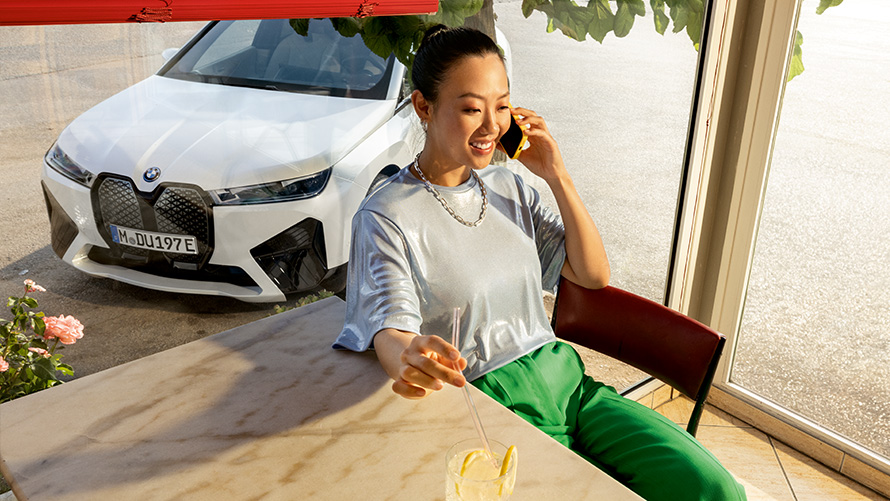 Conditions préalables pour Proactive Care Proactive Care, contrôle & entretien Conditions préalables Proactive Care Proactive Care, contrôle & entretien, femme assise à la table avec smartphone à la main et véhicule BMW en arrière-plan