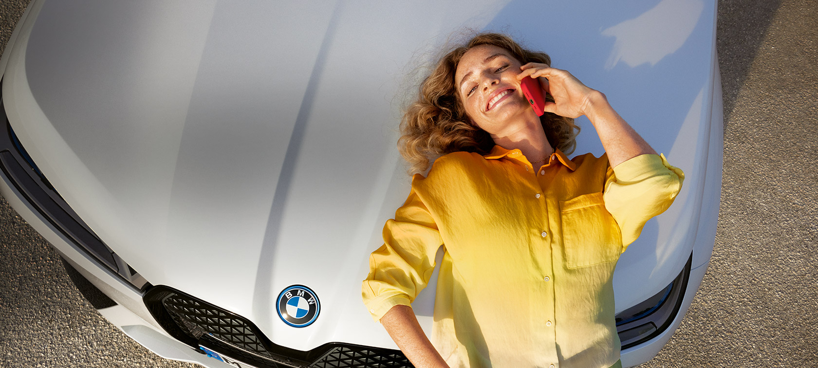 Relax We Care Proactive Care, contrôle & entretien Proactive Care, contrôle & entretien Relax We Care, femme avec chemise jaune et smartphone à la main adossée au capot BMW blanc